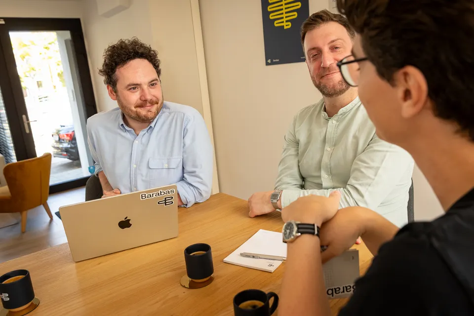 Twee Barabas-oprichters lachend in gesprek met een klant aan tafel, koffie erbij